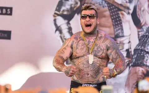 Popek kiedys, wytatuowany mężczyzna w okularach przeciwsłonecznych, z złotym łańcuchem i wisiorem, napina mięśnie.