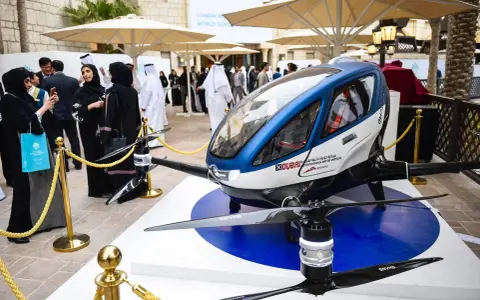 Ein futuristisches Flugtaxi, ein Dubai Drohne, steht auf einer Ausstellungsplattform. Menschen in traditioneller Kleidung betrachten das Gefährt.