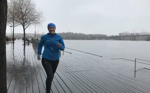 Glad kvinna med blå jacka och mössa springer i regn längs en träbrygga vid en sjö.