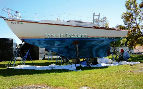 Un bateau est sur des chandelles, prêt pour une peinture. La ligne de flottaison est visible sur la coque bleue. Un homme travaille sur le bateau.