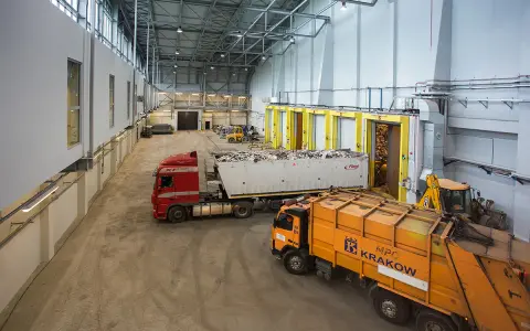 Wielka hala przemysłowa, gdzie trwa termiczne przekształcanie odpadów. Ciężarówki z odpadami czekają na rozładunek, a koparka przygotowuje materiał do przetworzenia.