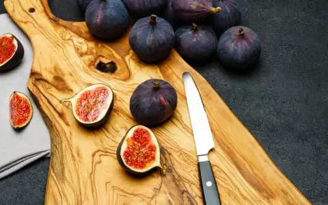 Kilka dojrzałych, ciemnofioletowych fig, niektóre przekrojone na pół, leżą na drewnianej desce obok noża.