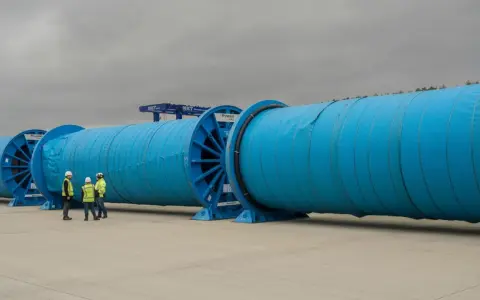 Große blaue Kabeltrommeln mit Arbeitern davor, Kräne im Hintergrund.