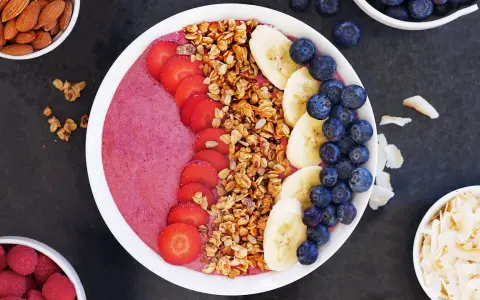 En färgglad acai bowl med jordgubbar, banan, blåbär och granola. Serverad med mandlar och kokosflingor vid sidan.