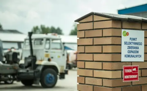 Tablica informuje, co to jest pszok – punkt selektywnego zbierania odpadów komunalnych. W tle widać ciężarówki.