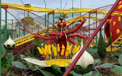 Eine rote Insektenfigur mit vielen Armen steht vor einer gelben Achterbahn.