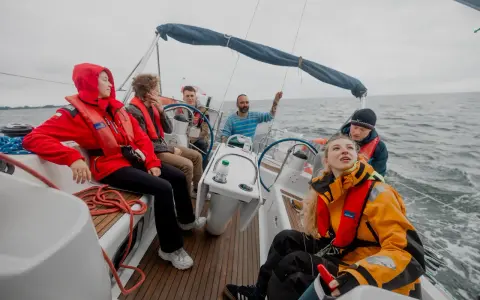 Grupa osób na jachcie, w kamizelkach ratunkowych, cieszy się rejsem. Jedna z osób trzyma ster, inni obserwują morze. Wszyscy wyglądają na zadowolonych, jakby właśnie zdobyli patent żeglarski.