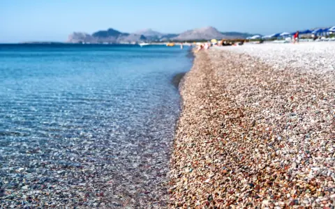 Rodos: turkusowe morze obmywa kamienistą plażę, w tle góry i plażowicze. Idealne miejsce na wakacje.