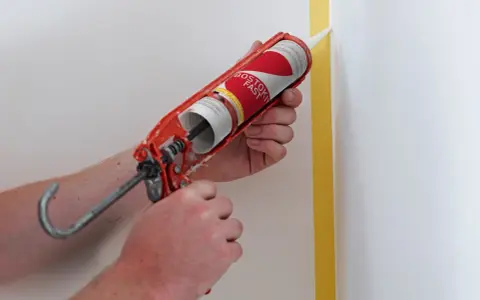Application de silicone avec un pistolet pour une finition parfaite. Le joint de silicone est appliqué le long d'un coin de mur masqué par du ruban de peintre.