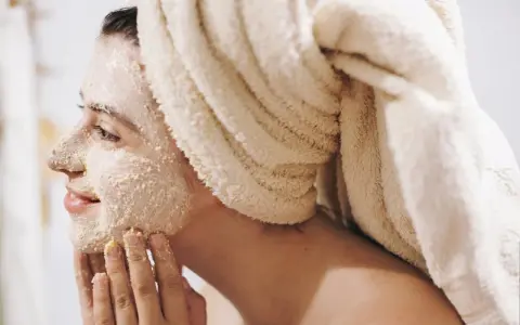 Kobieta z ręcznikiem na głowie nakłada na twarz peeling. To domowy sposób na gładką skórę.