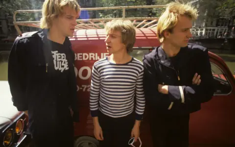 Le groupe Police, dans les années 80, pose devant un véhicule rouge. L'un porte un t-shirt à rayures, les autres des vestes sombres.