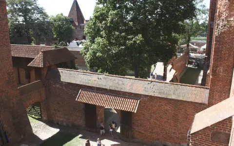 Wielki zamek w Malborku, ceglane mury, dziedziniec z drzewami i ludźmi spacerującymi pośród zabytkowej architektury.