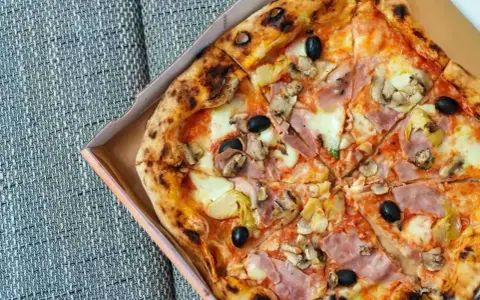 Pizza capricciosa z szynką, pieczarkami, karczochami i czarnymi oliwkami, w pudełku.