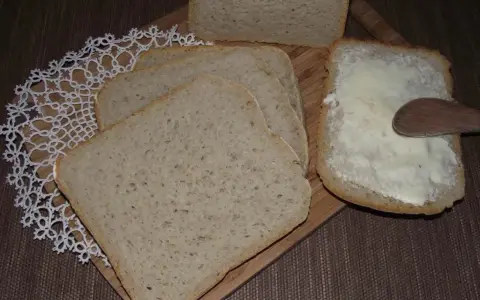 Chleb mieszany pokrojony w kromki, jedna z nich smarowana masłem drewnianą łyżką.