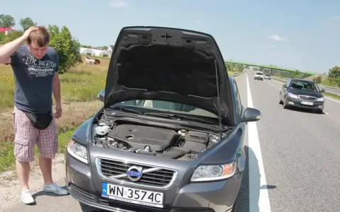 Mężczyzna zmartwiony przy otwartej masce Volvo, prawdopodobnie przegrzany silnik. W tle droga i pola.