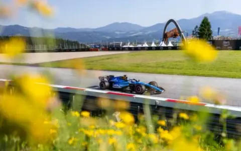 Un coche de F1 azul compite en un circuito soleado, con flores amarillas en primer plano. ¡Un gran resultado f1 hoy!