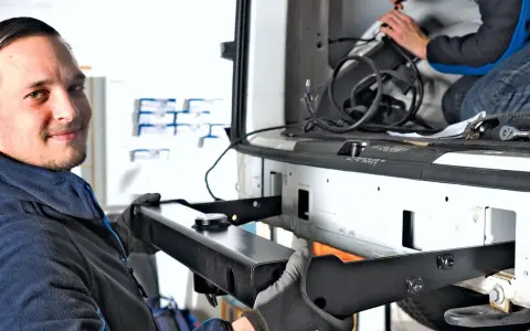 Mechanik z uśmiechem montuje hak w aucie. W tle widać drugą osobę pracującą przy instalacji.
