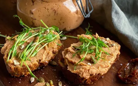 Två rostade brödskivor med krämigt refried beans, toppade med groddar och frön. En burk med pålägget syns i bakgrunden.