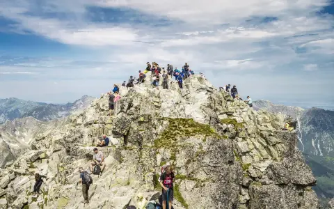 Tłum turystów na skalistym szczycie Rysów. Widać ludzi odpoczywających i podziwiających panoramę Tatr.