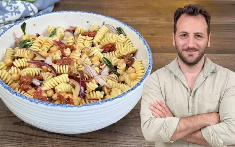 Un uomo sorride accanto a una ciotola di pasta fredda con ceci, pomodori secchi e cipolla rossa.