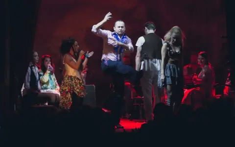 Scena teatralna, gdzie mężczyzna tańczy, a kobiety śpiewają. Atmosfera jak po pożarze w burdelu, pełna emocji i dramatyzmu.
