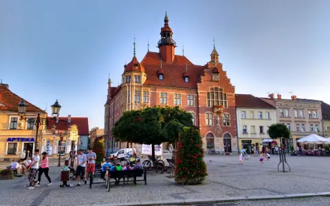 Rynek w Tarnowskich Górach tętni życiem. Ludzie spacerują, siedzą na ławkach, a dzieci bawią się na bruku. W tle widać zabytkowy ratusz.