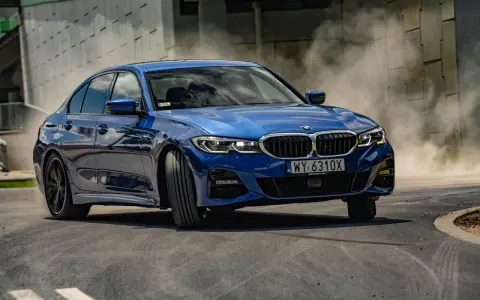 Niebieskie BMW 320i w zakręcie, unosząc tumany kurzu. Dynamiczna jazda na torze.