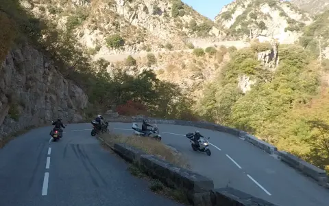 Motociclisti affrontano le curve del Col de Turini, immersi in un paesaggio montano roccioso e verdeggiante.