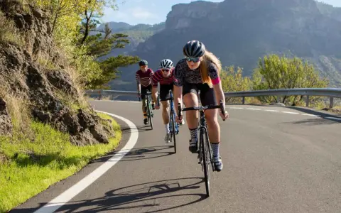 Three cyclists race up a mountain road. The lead cyclist, a woman, is what is a sportive person, focused and determined.