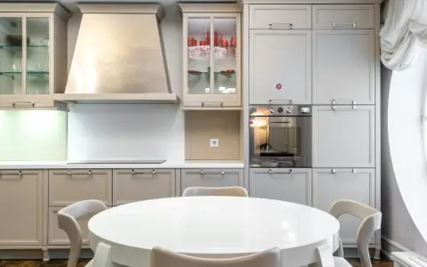 Cocina moderna con mesa redonda blanca y sillas. Gabinetes grises y beige, campana extractora de acero inoxidable. Ideal para ver catálogos de cocinas y precios.