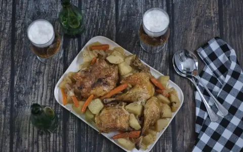 Pollo asado con patatas y zanahorias en una bandeja blanca, con cerveza y servilleta a cuadros.