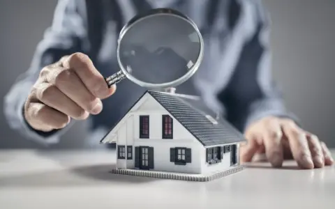Une main examine une maquette de maison avec une loupe, symbolisant l'analyse pour un crédit construction maison.