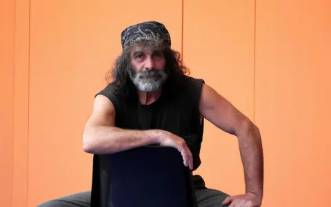 Mauro Corona, con bandana e barba grigia, seduto su una sedia. Immagina i suoi libri in ordine cronologico.