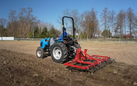 Niebieski traktor z czerwoną broną wykonuje bronowanie pola. W tle drzewa i błękitne niebo.