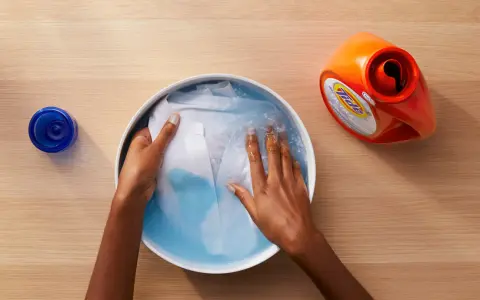 Manos lavando ropa blanca en un bol con agua y detergente. A un lado, un bote de detergente y un tapón azul.
