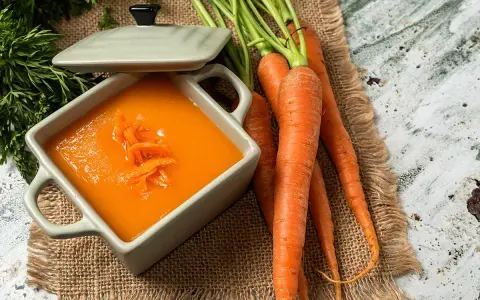 Un tazón de cremoso puré de zanahoria, adornado con hebras de zanahoria rallada, junto a zanahorias frescas y verdes.