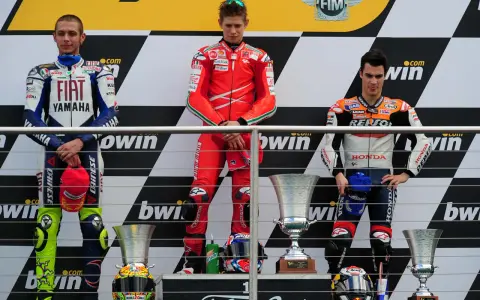 Podio con tre motociclisti famosi in tuta: Rossi, Stoner e Lorenzo. Vittoria celebrata con trofei.