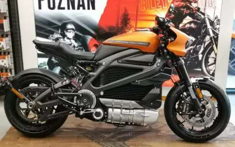 Pomarańczowy harley elektryczny z czarnymi akcentami, prezentowany na drewnianej podłodze, z plakatem Harley-Davidson w tle.