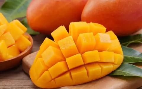 Dojrzałe mango, pokrojone w kostkę, prezentuje swoje soczyste, pomarańczowe wnętrze. Obok leżą całe owoce i liście.