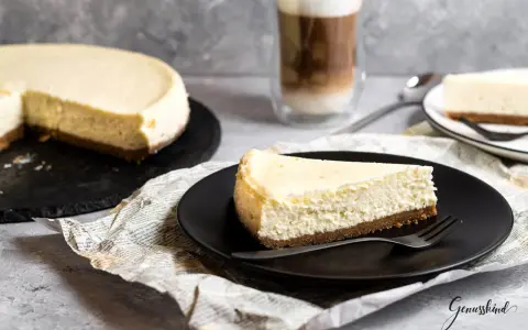 Ein Stück New York Cheesecake auf schwarzer Platte, daneben ein ganzer Kuchen und ein Latte Macchiato.