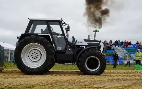 Ciężki traktor Ursus 1224 z potężnym dymem z rury wydechowej podczas zawodów tractor pulling. Widownia obserwuje z trybun.