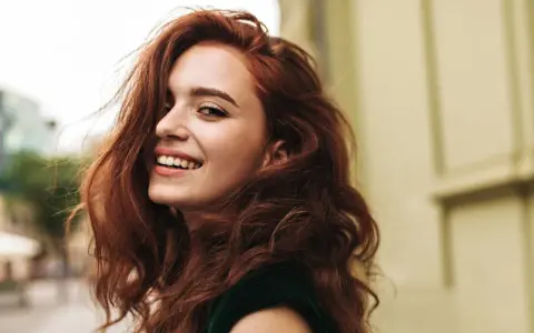 Donna con capelli rossi mossi e un sorriso radioso. La sua chioma è così bella che si può fare la tinta ogni 15 giorni per mantenerla perfetta.