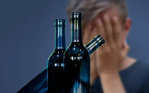 Trzy butelki alkoholu na pierwszym planie, w tle osoba zakrywająca twarz. Symbol walki z nałogiem, nadzieja na odwyk alkoholowy.