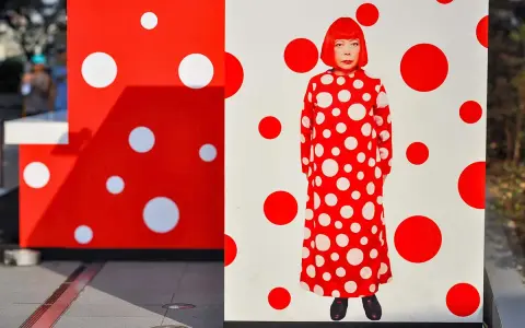 A woman in a red and white polka dot dress stands amidst Yayoi Kusama patterns.