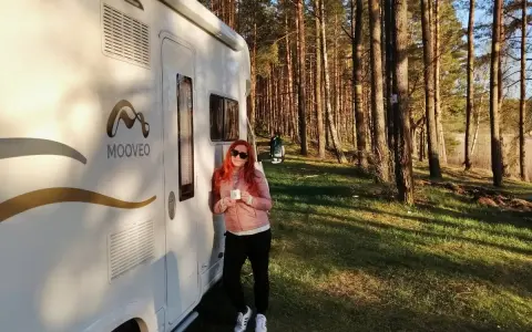 Kobieta z kubkiem kawy opiera się o kampera na leśnej polanie. Wspaniała przygoda w **Finlandii kamperem**!