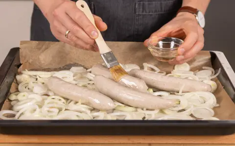 Bratwurst im Backofen: Zwiebelringe auf Backpapier, darauf vier rohe Bratwürste. Mit einem Pinsel wird Öl aufgetragen.