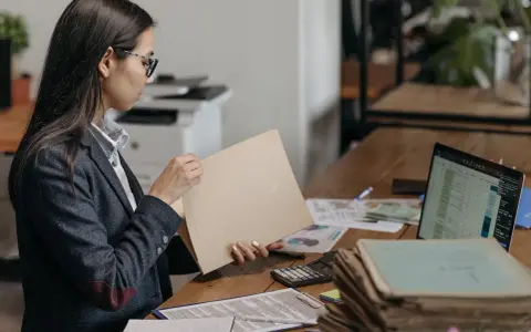 Kobieta w okularach przegląda dokumenty, szukając tytułu prawnego do lokalu. Na biurku leżą też laptop, kalkulator i sterty papierów.
