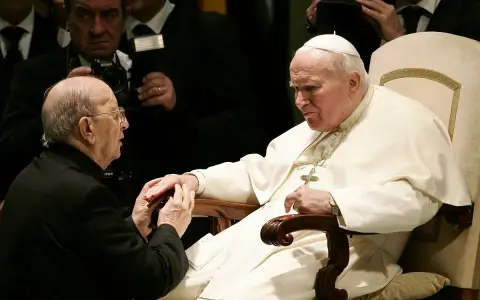Papież Jan Paweł II w białej szacie, klęczący przed nim mężczyzna w czerni. W tle widać innych duchownych i dziennikarzy, być może z zakonu **jezuitów**.