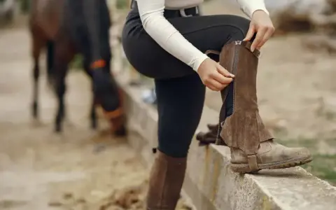 Jeździec zapina brązowe sztylpy na zamek, gotowy do jazdy. W tle widać konia.