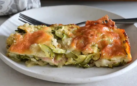 Una fetta di sformato di zucchine dorato e filante, con formaggio fuso e zucchine a rondelle, servito su un piatto bianco.